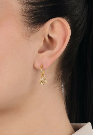 Goldene Creolenohrringe mit einem herzförmigen Anhänger, der ein filigranes Knotendesign aufweist, hängen unterhalb des Ohrläppchens auf einem glatten, hellen Teint.