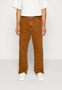 Jean en denim marron avec une coupe droite, doté d'une fermeture à boutons et de passants pour ceinture. Porté avec des baskets blanches.