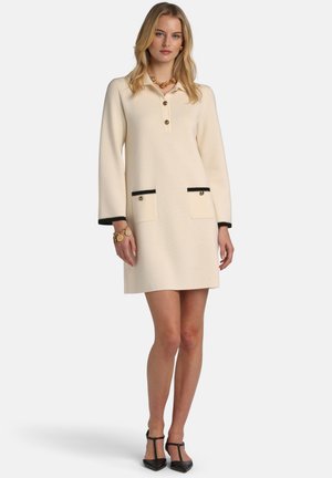 Mujer vestida con un vestido crema hasta la rodilla con bolsillos con ribete negro, botones dorados y tacones negros con tira en T, de pie contra un fondo sencillo.