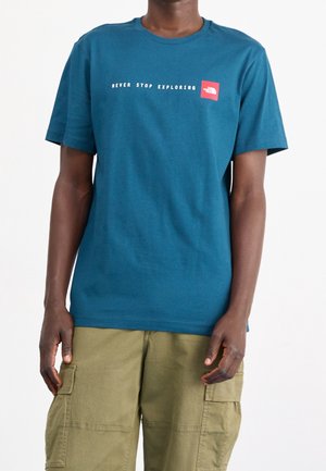 Teal katoenen t-shirt met een witte tekstgrafiek "NEVER STOP EXPLORING" en een rood vierkant logo op de linkerborst. Korte mouwen, ontspannen pasvorm.