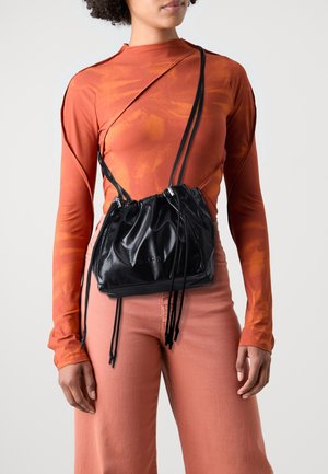 Svart lær drapepose med samlet tekstur og lange stropper, kombinert med en oransje mønstret langermet topp og rosa vide bukser.