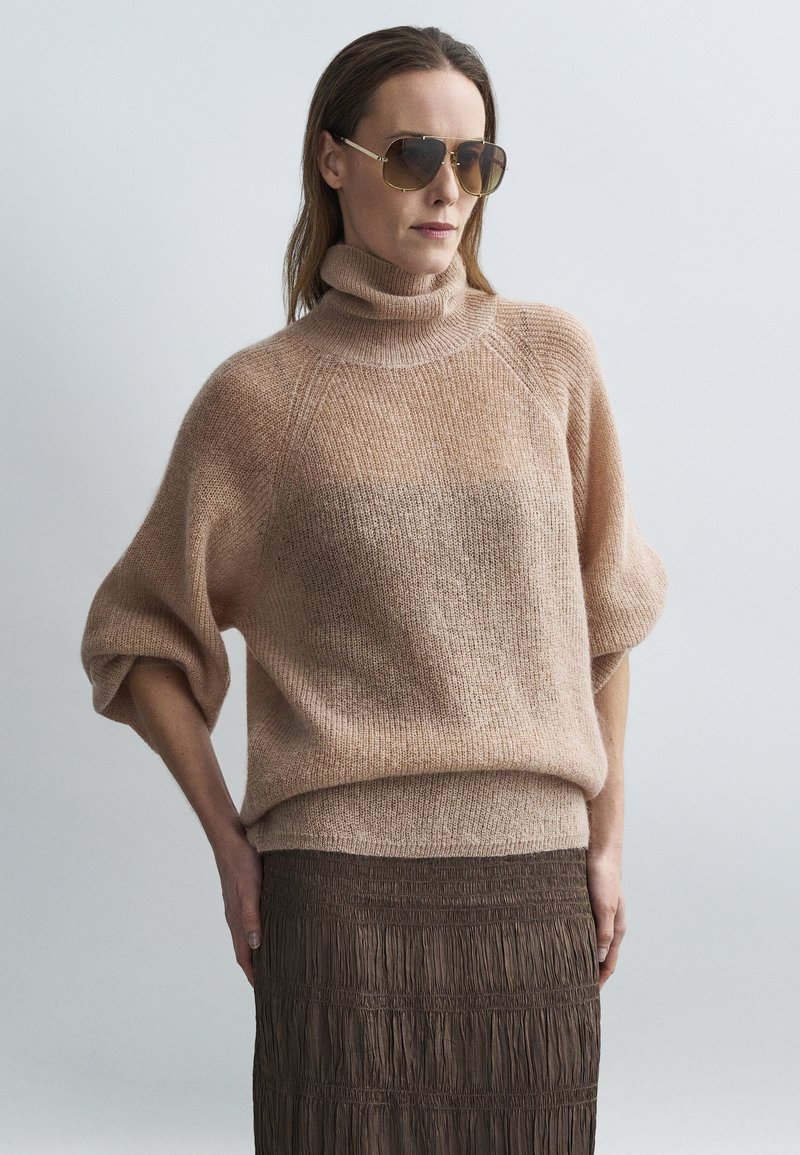 Nagy méretű bézs kötött pulóver magas gallérral és rövid ujjakkal. Sötétbarna, texturált, ráncolt szoknyával párosítva.