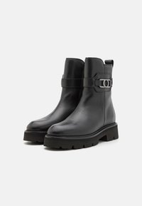 Bottines en cuir noir avec un bout rond, une semelle en caoutchouc épaisse et une sangle dotée d'un accent métallique circulaire. Texture lisse.