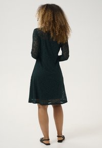 Lace dress in deep green with long sleeves, featuring a fitted bodice and flared skirt. Subtle floral pattern and scalloped hem detail.