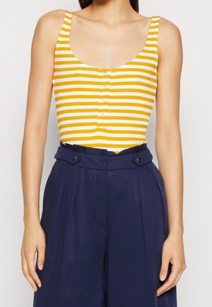 Vrouw draagt een mouwloze gele en witte gestreepte top met knopen, ingestopt in hoge taille marineblauwe geplooide broek met knopdetails.