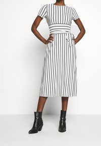 Vit klänning med svarta vertikala ränder, korta ärmar, en åtsittande midja och en knytDetalj framtill, tillsammans med svarta ankelstövlar.