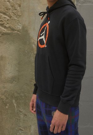 Person wearing a black hoodie with an orange and white basketball logo, paired with blue and green plaid pants, standing against a textured wall.