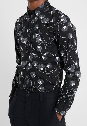 Man draagt een zwart overhemd met witte en grijze abstracte bloemenprint, ingestopt in een zwarte broek met handen in de zakken.