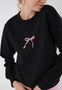 Svart hettegenser laget av mykt materiale, med ribbede mansjetter og kant, og med en liten rosa sløyfe brodert på midten av fronten.