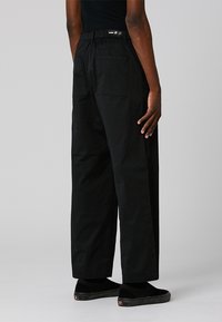 Personne portant un pantalon noir ample avec des poches arrière et des chaussures noires sans lacets, se tenant devant un fond gris uni.