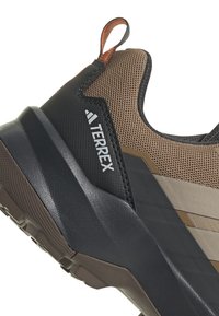 Brązowe i czarne buty sportowe z teksturowaną cholewką, z wyraźnym logo, amortyzowaną podeszwą oraz pętlą na zapięcie ułatwiająca zakładanie.