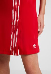 Röd atletisk klänning med vit tre-stripesdesign, logotyp vid fållen och en sidoklyfta för rörelsefrihet. Tillverkad av ett mjukt, stretchigt material.