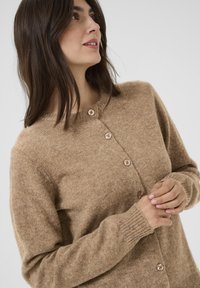 Pletený cardigan v měkké béžové textuře s kulatým výstřihem, pěti dřevěnými knoflíky a žebrovanými manžetami, který nabízí volný střih.