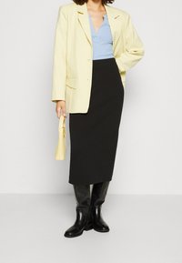 Hellgelbe Blazer über einem blauen gerippten Oberteil, kombiniert mit einem schwarzen Bleistiftrock und kniehohen schwarzen Stiefeln. Hält eine kleine gelbe Tasche.