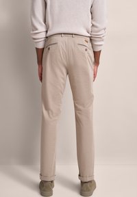 Person in beigen Chinos, beigem geripptem Pullover und olivgrünen Wildlederschuhen, der mit dem Rücken zu einem hellbeigen Hintergrund steht.