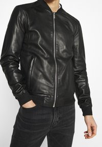 Svart läderbombarjacka med ribbad krage, framdragkedja och sidofickor. Har en slät textur och åtsittande ärmar.