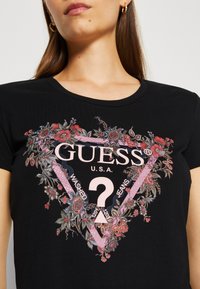 černé tričko s květinovou trojúhelníkovou grafikou. Design obsahuje text "GUESS U.S.A" a "WASHED JEANS" v růžové barvě.