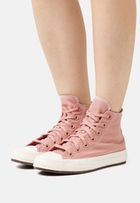 Legs wearing pink high-top canvas sneakers with white rubber soles and toe caps, standing on a white background.