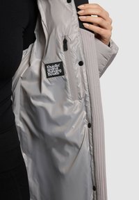 Cappotto imbottito grigio chiaro con design trapuntato. L'interno presenta un'etichetta con il logo nera e una tasca con zip. Materiale liscio e lucido.