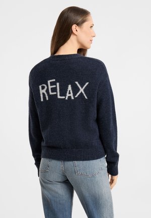 Frau mit langen braunen Haaren, die einen marineblauen Pullover mit dem Wort "RELAX" in Weiß auf dem Rücken und hellblaue Jeans trägt, von hinten.