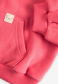 Manche et poche en sweat-shirt polaire rose avec poignet côtelé et petite étiquette arborant un logo simple en forme de visage d'ourson.