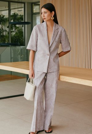 Femme portant un costume gris clair à motifs avec des manches courtes et un pantalon à jambes larges, tenant un sac tissé, debout dans une pièce moderne avec une table en bois.