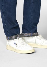 Baskets en cuir blanc avec un bout perforé, semelle beige et lacets décontractés, associées à un jean bleu foncé avec un rabat de doublure à carreaux.