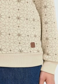 Sudadera beige claro con un patrón geométrico marrón, que presenta una cintura y puños acanalados. Incluye una pequeña etiqueta de marca marrón en el lado.