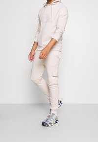 Felpa beige con design grafico, abbinata a pantaloni cargo coordinati e scarpe sportive grigie con accenti colorati e suole strutturate.