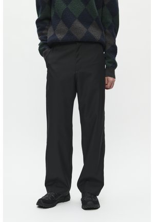 Pantaloni negri, cu croială liberă și textură netedă. Combinat cu adidași negri. Modelul stă cu mâinile în buzunare.