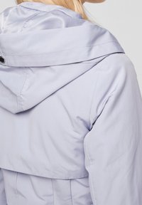 Veste lavande claire avec capuche, texture lisse et détails de coutures visibles. Présente une coupe décontractée et un design de capuche pratique.