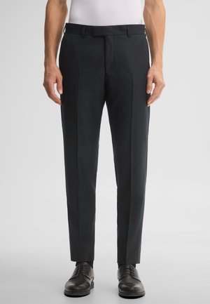 Uomo che indossa pantaloni da abito neri su misura, scarpe eleganti nere e una camicia bianca infilata, in piedi davanti a uno sfondo chiaro e uniforme.