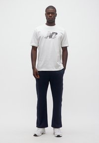 Άνδρας με λευκό κοντομάνικο μπλουζάκι με σχέδιο, σκούρο παντελόνι και λευκά αθλητικά παπούτσια, στέκεται μπροστά σε ένα απλό ανοιχτό γκρι φόντο.