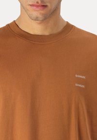 Barna pamut póló, kerek nyakkivágással. Két fehér "SAM SøE" logó található a bal oldalon. Sima textúrájú, egyszerű dizájnnal.