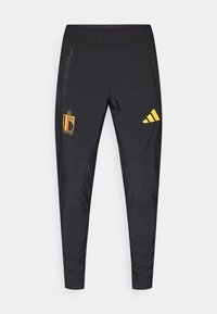 Čierne športové nohavice s vreckami na zips, s žltým belgičanským emblémom a žltým logom Adidas na ľavom stehne. Hladký povrch.