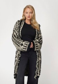 Karirane čierno-biele sako, mäkká textúra, dlhé rukávy s strapcami, otvorený predok, nosené cez čierny top a tmavé nohavice.