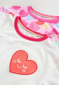 Dve bombažni majici: ena bela z rdečo srčno nalepko in ena roza s pisanimi vzorci src ter mavričnimi poudarki.