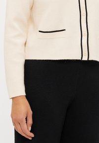 Krems krāsas kardigāns ar melnu apdari, ar pogām un kabatu; mīksta tekstūra ar īsu dizainu virs melnām biksēm.