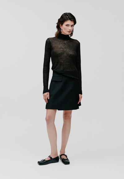 Haut noir à manches longues avec une finition transparente et texturée, associé à une jupe noire en A unie dotée d'une poche latérale, portée avec des chaussures plates noires.