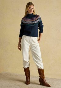 Marineblauer Pullover mit buntem Fair-Isle-Muster, weiße Jeans mit lockerem Sitz und braune Cowboy-Stiefel mit detailliertem Design.