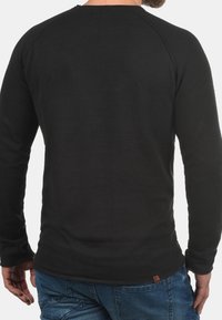 Chemise noire à manches longues avec une texture côtelée et un col rond. Présente des manches raglan et une petite étiquette marron sur l'ourlet inférieur.