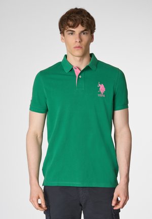 Giovane uomo che indossa una polo verde con colletto rosa e un logo rosa, in piedi davanti a uno sfondo bianco semplice.