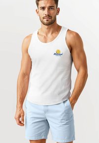 White cotton tank top with blue embroidered logo and lemon detail, paired with light blue shorts, featuring side pockets and smooth texture.