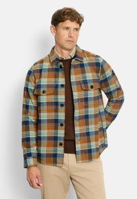 Karierte Jacke in Braun, Blau und Türkis; mit durchgehender Knopfleiste, zwei oberen Taschen und lässigem Schnitt. Trägt man über einem braunen Pullover.