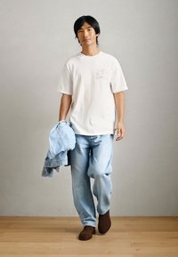 Λευκό κοντομάνικο μπλουζάκι με γραφικό, ανοιχτό μπλε τζιν και ελαφριά τζην μπουφάν κρεμασμένο στον βραχίονα. Καφέ υποδήματα ολοκληρώνουν το σύνολο.