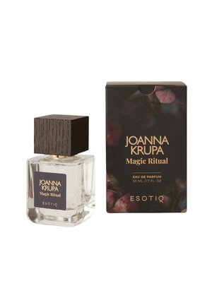 Przezroczysta szklana butelka perfum z ciemnym drewnianym korkiem, oznaczona etykietą "Joanna Krupa Magic Ritual", obok pasującego czarnego pudełka z kwiatowymi akcentami.