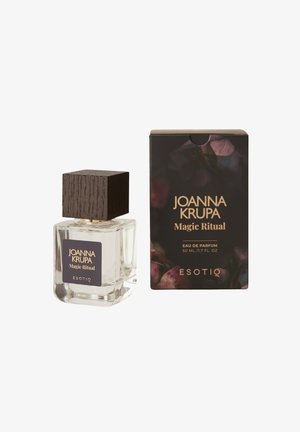 Przezroczysta szklana butelka perfum z ciemnym drewnianym korkiem, oznaczona etykietą "Joanna Krupa Magic Ritual", obok pasującego czarnego pudełka z kwiatowymi akcentami.