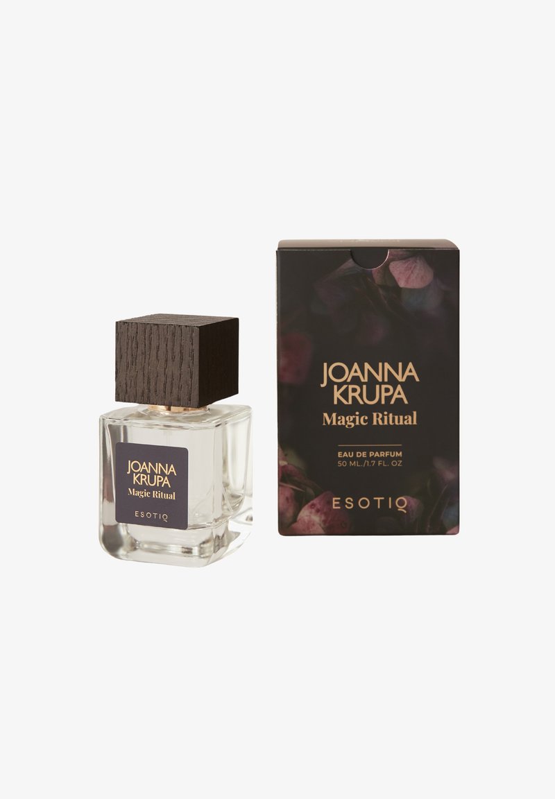 Przezroczysta szklana butelka perfum z ciemnym drewnianym korkiem, oznaczona etykietą "Joanna Krupa Magic Ritual", obok pasującego czarnego pudełka z kwiatowymi akcentami.