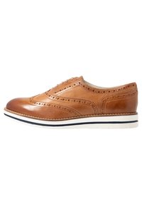 Chaussure brogue en cuir tan avec des perforations décoratives, bout arrondi et semelle rayée blanche et marine. Présente des détails de couture contrastés.