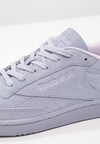 Basket en suède violet clair avec bout rond, surface texturée et lacets plats. Présente un logo en relief et des détails de couture minimalistes sur le côté.